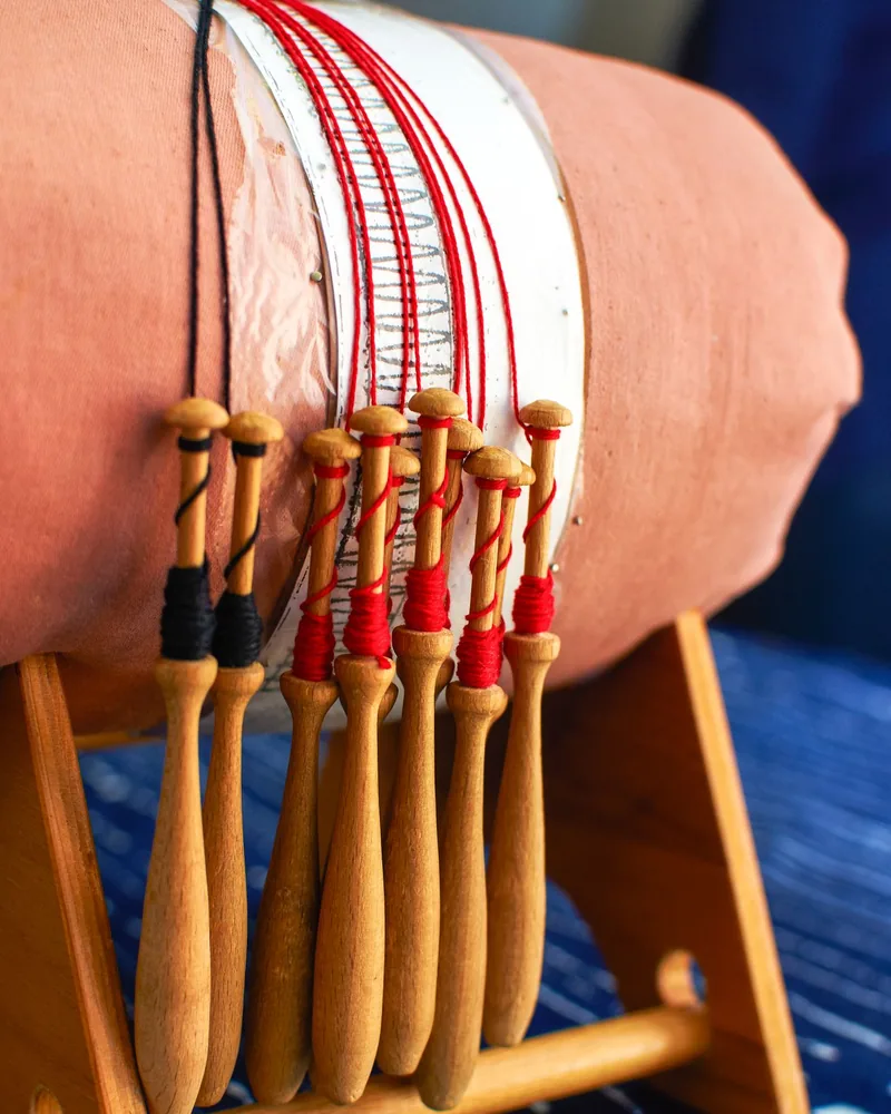 Bolillos de madera con hilo rojo y negro colgando de un mundillo cilíndrico sujeto en su soporte